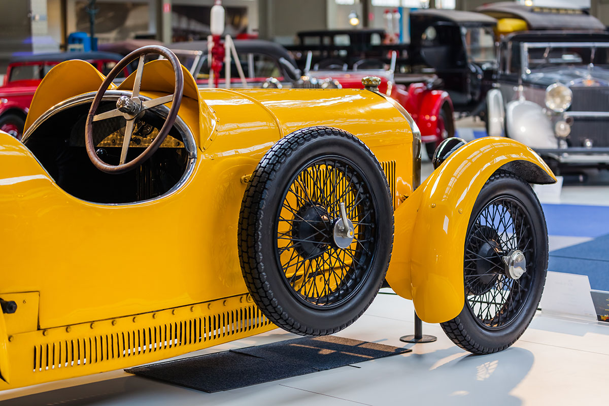 A vintage mustard yellow open top car with black wheels and a metal steering wheel in a museum surrounded by other vintage vehicles